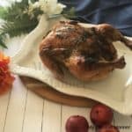 White platter with a whole roasted chicken on it. The platter is on a wood board with a blue napkin behind it. There are flower next to the napkin and apples in front of the platter.