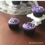 Cupcakes with purple frosting on a white cupcake stand and some on a glass cake stand in the back ground.