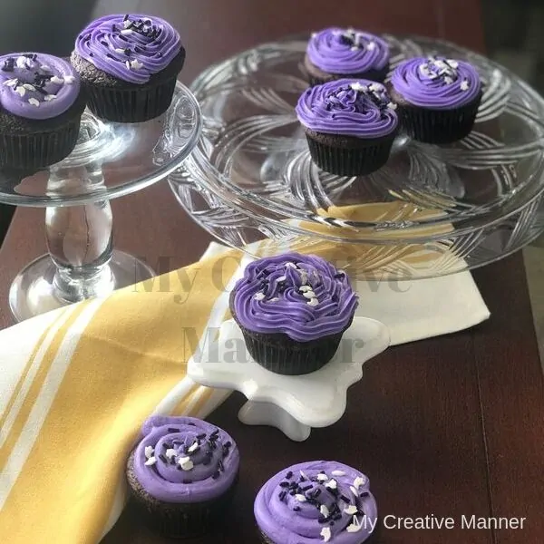 White and yellow napkin is under cupcakes that have purple frosting. Some cupcakes are on a white cupcake stand while others are on two glass cake stands.