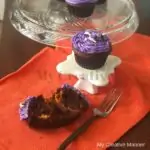 Halloween cupcakes with purple frosting on a orange napkin. Some cupcakes on a glass cake stand. There is a fork on the napkin along with cupcake liners.