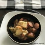 Black bowl with kielbasa, sauerkraut and potatoes in it. The bowl is sitting on a white plate that has a black charger under it. A black and white plaid check napkin is behind it with a fork on it.
