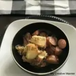 Black bowl with kielbasa, sauerkraut and potatoes in it. The bowl is sitting on a white plate that has a black charger under it. A black and white plaid check napkin is behind it with a fork on it.