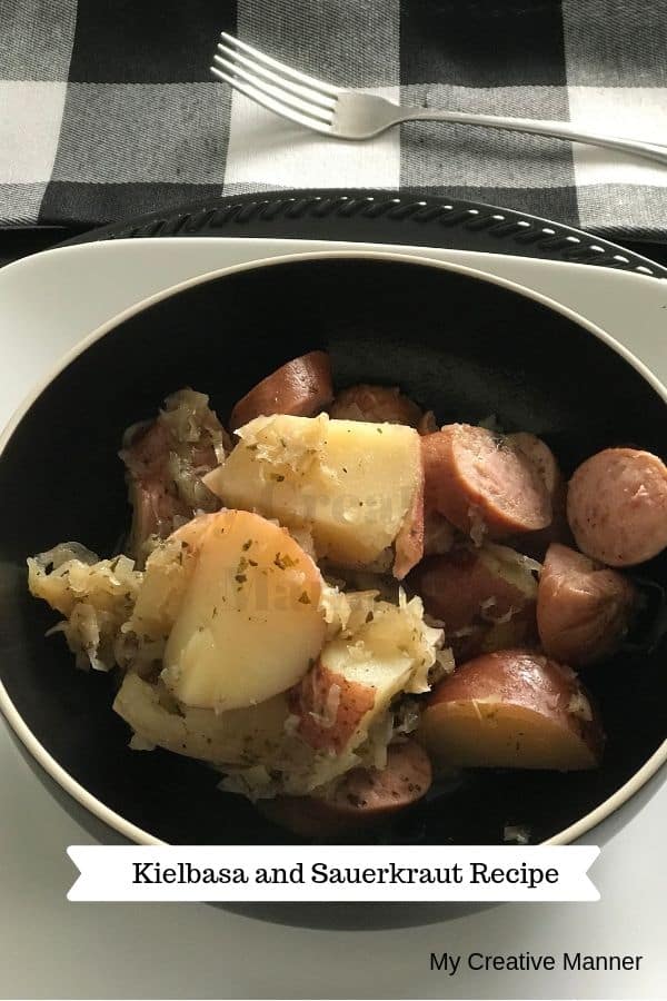 Black bowl with kielbasa, sauerkraut and potatoes in it. The bowl is sitting on a white plate that has a black charger under it. A black and white plaid check napkin is behind it with a fork on it.