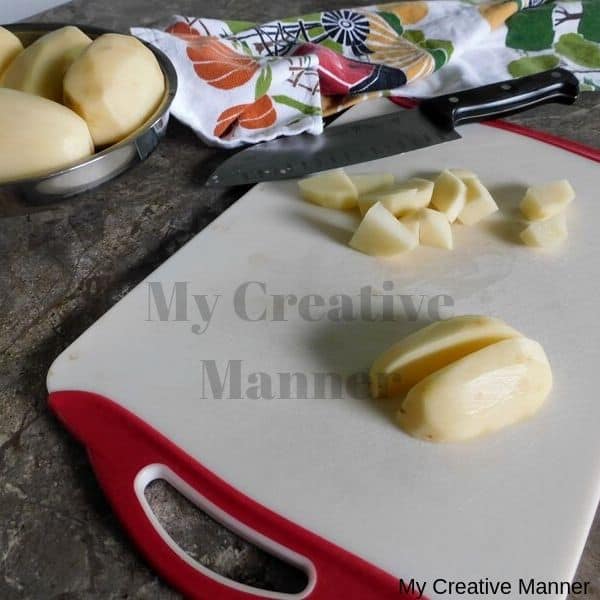 Cutting board with a potato that has been cut in half and is diced up. There is also a knife on the cutting boards and behind is a lod with more potatoes in it.