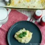 Grey plate with loaded mashed potatoes on it. Red salt and pepper shakers are behind the plate. The plate is sitting on a red and white napkin with a glass of milk and a baking dish full of the potatoes.