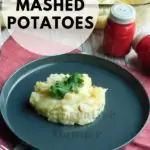 Grey plate with loaded mashed potatoes on it. Red salt and pepper shakers are behind the plate. The plate is sitting on a red and white napkin with a glass of milk and a baking dish full of the potatoes.