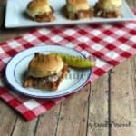 Red and white napkin under a white and blue plate. The plate has a BBQ chicken slider and pickle on it.Behind the plate is a white plate with three more sliders on it.
