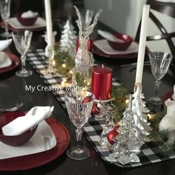 Black and white table runner with clear glass Christmas decorations and red candles. Red charger plates with a white plate and red bowl on them. There is a white napkin in each red bowl. Crystal glasses and silverware adron each place setting.