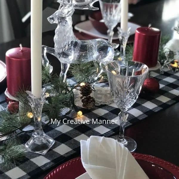 Black and white table runner with clear glass Christmas decorations and red candles. Red charger plates with a white plate and red bowl on them. There is a white napkin in each red bowl. Crystal glasses and silverware adron each place setting.