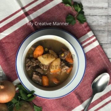 A white and blue bowl on a red and white napkin. The bowl is filled with Instant Pot beef stew with vegetables. There is a spoon on the napkin as well as an onion and fresh parsley.