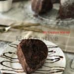 A piece of chocolate cake on a white plate that has chocolate sauce on it. In the back ground you can see a fork and knife and the rest of the bundt cake.