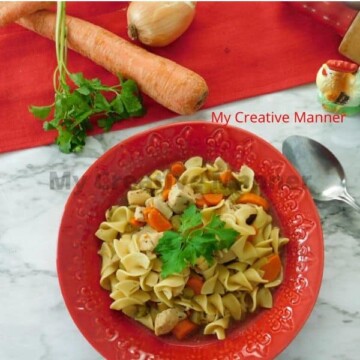 Red bowl filled with pressure cooker chicken noodle soup. In the background is a red towles, a carrot, fresh parsley, an onion, and a roaster figure.