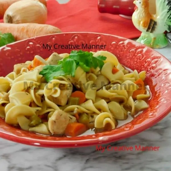 Red bowl filled with pressure cooker chicken noodle soup. In the background is a red towles, a carrot, fresh parsley, an onion, and a roaster figure.