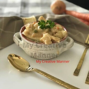 A bowl filled with Instant Pot chicken and dumplings. There is a spoon in front of the bowl and next to the bowl is a fork and knife. Behind the bowl is an onion and carrot.