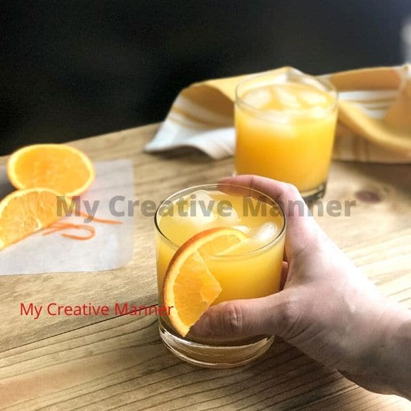 Two glasses that have fuzzy navel in it. There is a hand that is holding one of the glasses. with an orange that is cut up next to them on wax paper.