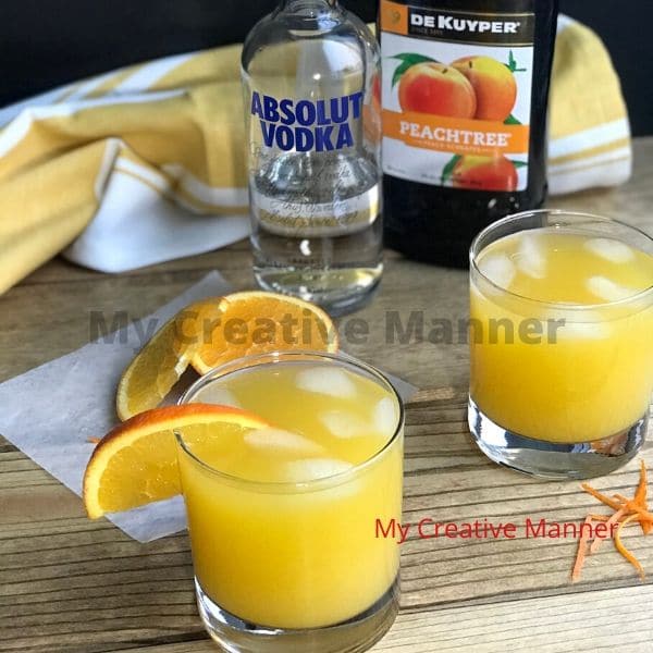 Two glasses of filled with fuzzy navel cocktail. A bottle of peach schnapps and vodka are in the background with a yellow and white towel.