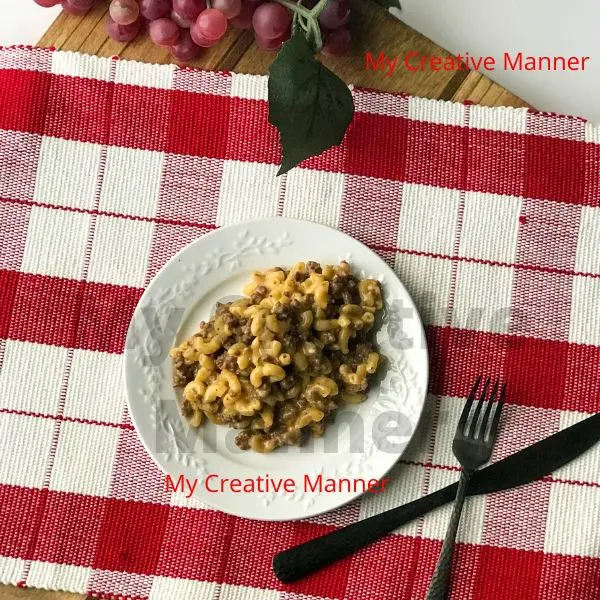 A white plate that has hamburger macaroni on it. The plate is sitting on a red and white placemat. Next to the plate is a fork and knife. Above the plate is grapes.