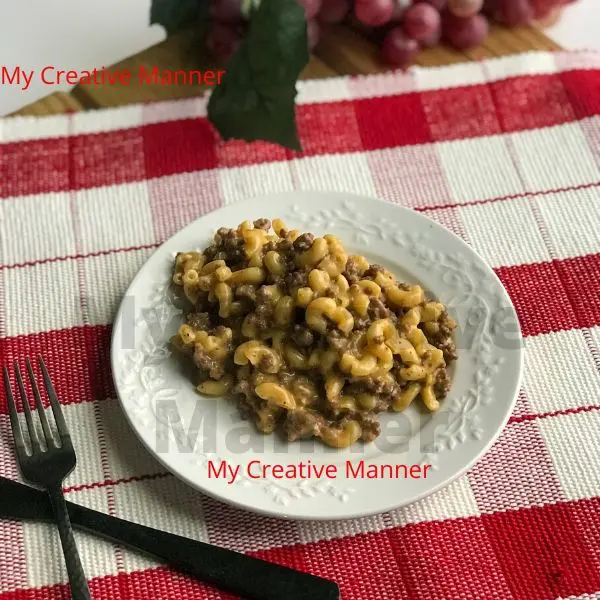 A white plate that has hamburger macaroni on it. The plate is sitting on a red and white placemat. Next to the plate is a fork and knife. Above the plate is grapes.