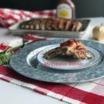 A white and blue plate that has two pork ribs on it. The plate is sitting on a metal charger and a red and white placemat. In the background there is a fork, red and white napkin, bottle of bbq sauce, and a cow and pig pepper and salt shaker.