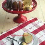 Lemon pound cake recipe on a cake stand with a piece of it in front on a white plate.