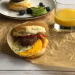 Breakfast biscuit that is wrapped and one on a plate. A glass of orange juice is next to it.