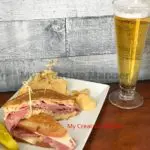 A grilled sandwich on a plate with chips and a pickle, next to it is a beer.