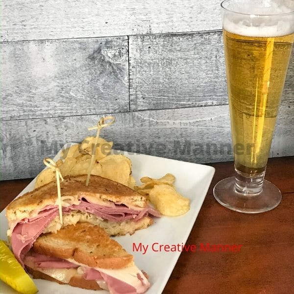 A grilled sandwich on a plate with chips and a pickle, next to it is a beer.