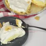 Slice of banana cream pie with a graham cracker crust on a plate with a fork on the plate. The whole pie is next to the plate.