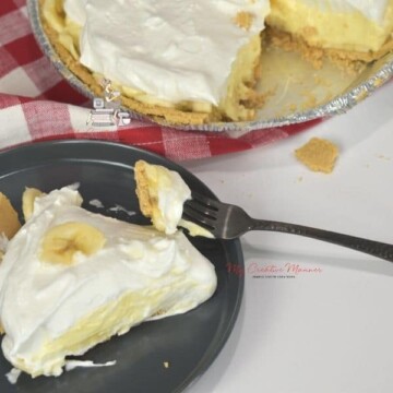 Slice of banana cream pie with a graham cracker crust on a plate with a fork on the plate. The whole pie is next to the plate.