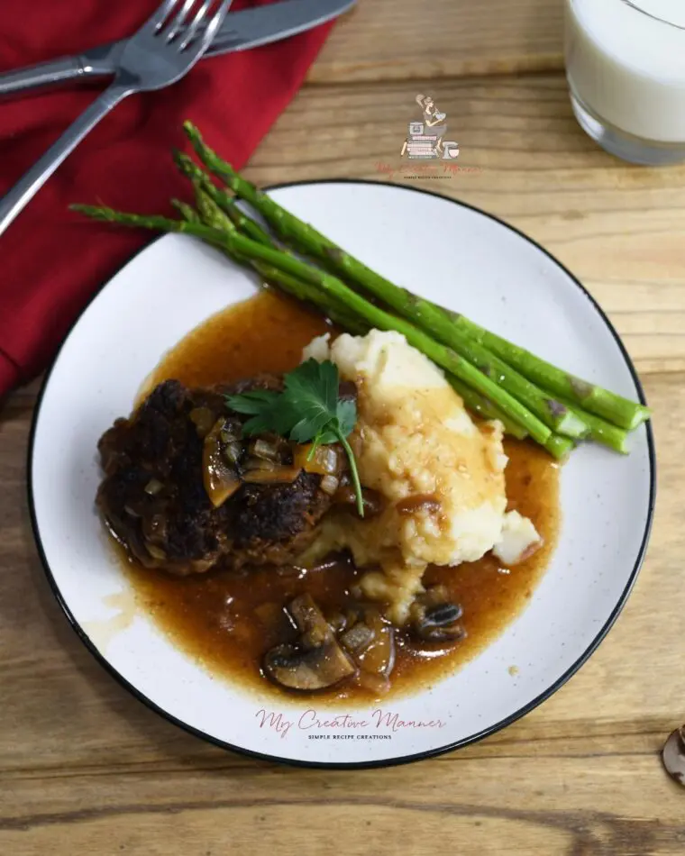 Hamburger steak With Gravy that is covering mashed potatoes. Along with asparagus on a plate.