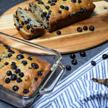 Two loaves of fresh baked blueberry banana bread. One on a cutting board and the other in a baking dish.