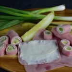 Lunch meat ham with cream cheese on it. Pickles and green onions are on a wood board with some completed polish roses and pickle wraps.