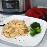 Slow cooker chicken and gravy over mashed potatoes on a white plate with broccoli next to it.
