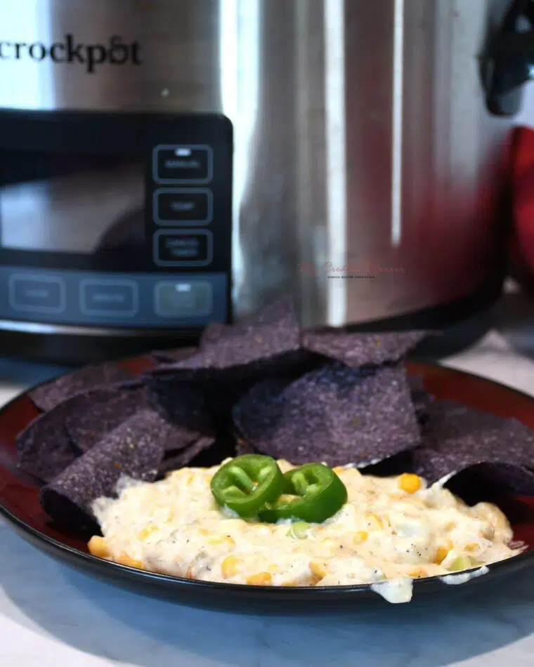 A slow cooker in the background with a plate that has chips and corn dip on it.