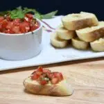An antipasto recipe for classic bruschetta recipe in front of a plate that has a bowl filled with the tomato bruschetta. And garlic toast is next to it.