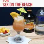 A bottle of vodka and peach schnapps is behind a hurricane glass that is filled with sex on the beach cocktail.
