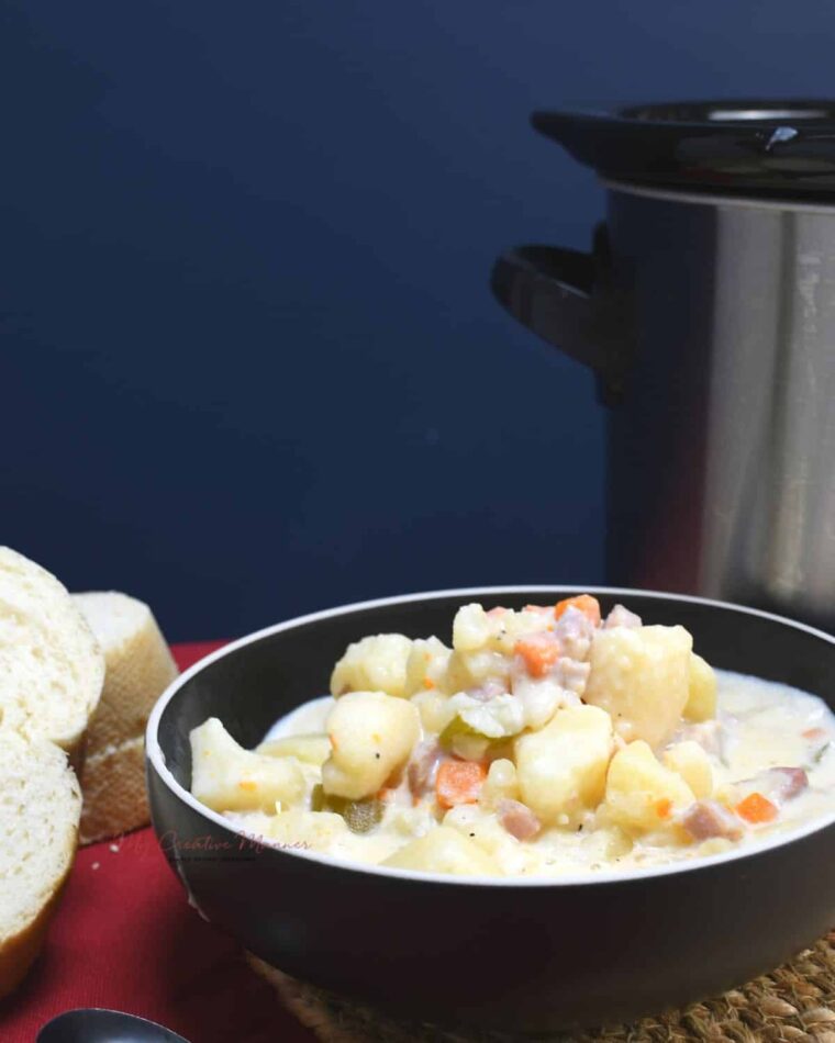 A bowl filled with slow cooker potato and ham soup with a crockpot next to the bowl.