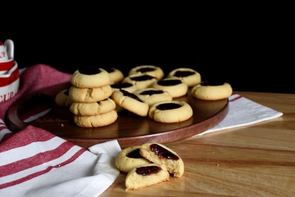 Two thumbprint cookies in front of a board with more raspberry thumbprint cookies on it.