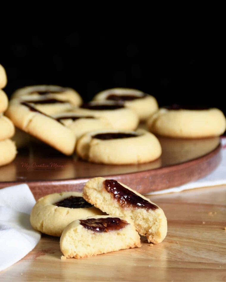 A close up of a raspberry thumbprint cookie that is broke in half. More of the raspberry jam filled cookies are in the back ground.