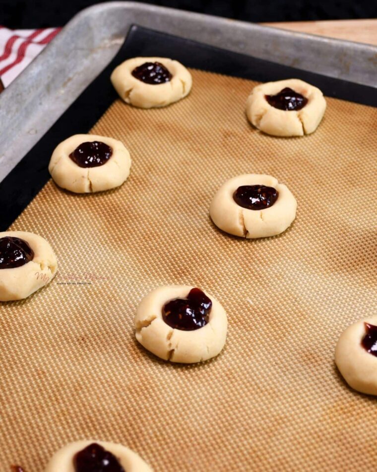 Unbaked thumbprint cookies that are filled with raspberry jam on a baking sheet pan.