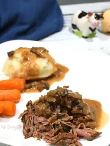 A close up of crockpot roast that is covered with a creamy mushroom gravy on a plate with mashed potatoes and carrots.