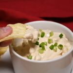 A chip that is being held that has onion dip with more of the 3 ingredient dip in a bowl.