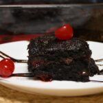 A close up of a 3 ingredient chocolate cherry brownie on a plate that is topped with a cherry.