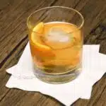 A rocks glass filled with a classic old fashion cocktail with a large ice cube and an orange peel.