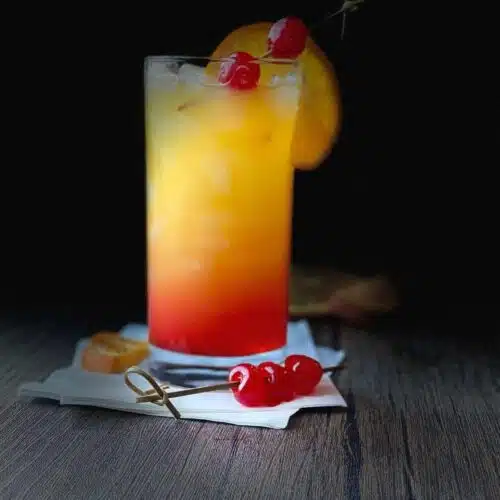 A highball class filled with tequilla sunrise and garnished with an orange slice and maraschino cherries.