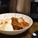 A bowl filled with ice cream and apple cobbler with a crockpot behind the bowl.