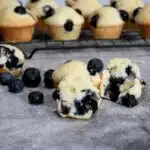 Close up of a mini blueberry muffin that is split in half with fresh blueberries next to it. And more mini muffins on a cooling rack.