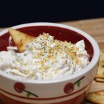 A bowl filled with the everything bagel dip recipe and a pita chip is in the dip.
