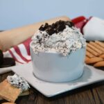 A bowl filled with oreo dip with a graham cracker that has some on it.