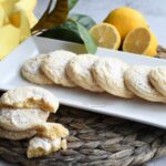 A plate that has the lemon drop cookies on it with a stack of them in front of the plate.